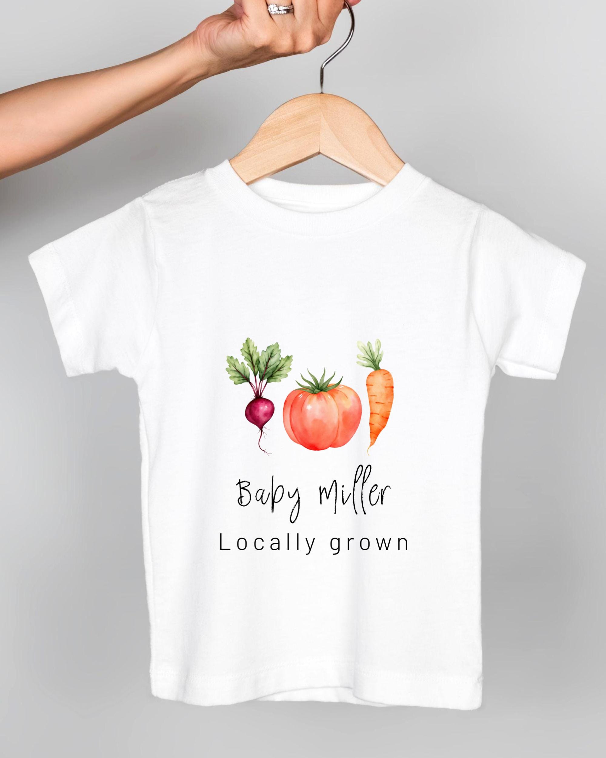 A white t-shirt with a graphic design of a carrot, tomato, and radish, along with the text "Baby Miller Locally grown".