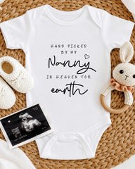 A white baby onesie with the text "Hand picked by my Nanny in Heaven for earth" printed on it, along with a crocheted bunny toy and a black and white photograph of an infant's head.