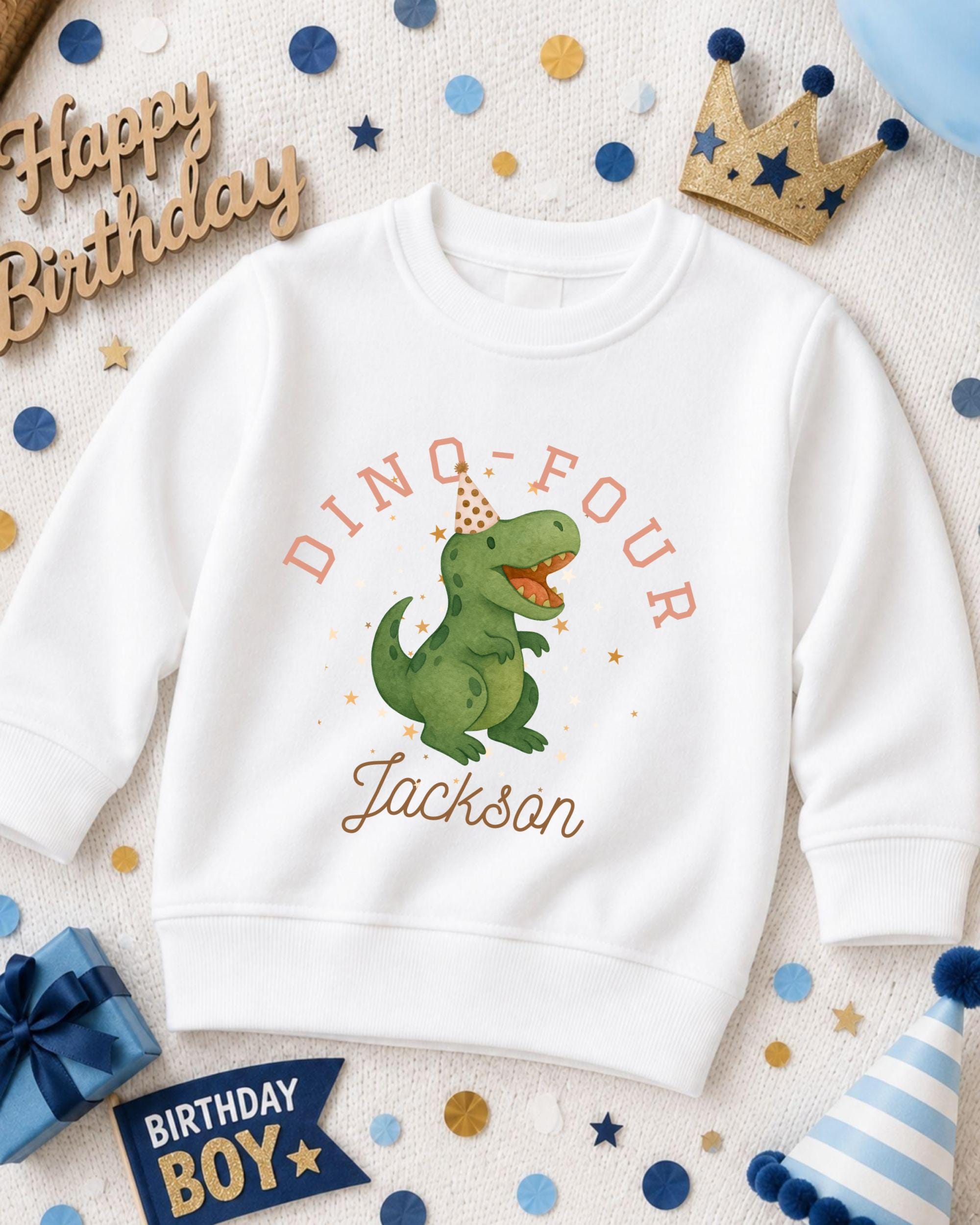 A white sweatshirt with a dinosaur graphic and the text "DINO FOUR JACKSON" printed on it, surrounded by birthday decorations such as a crown, confetti, and a birthday banner.