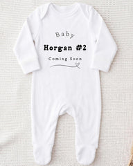 A white onesie with text printed on it, indicating that the baby is expected to arrive soon.