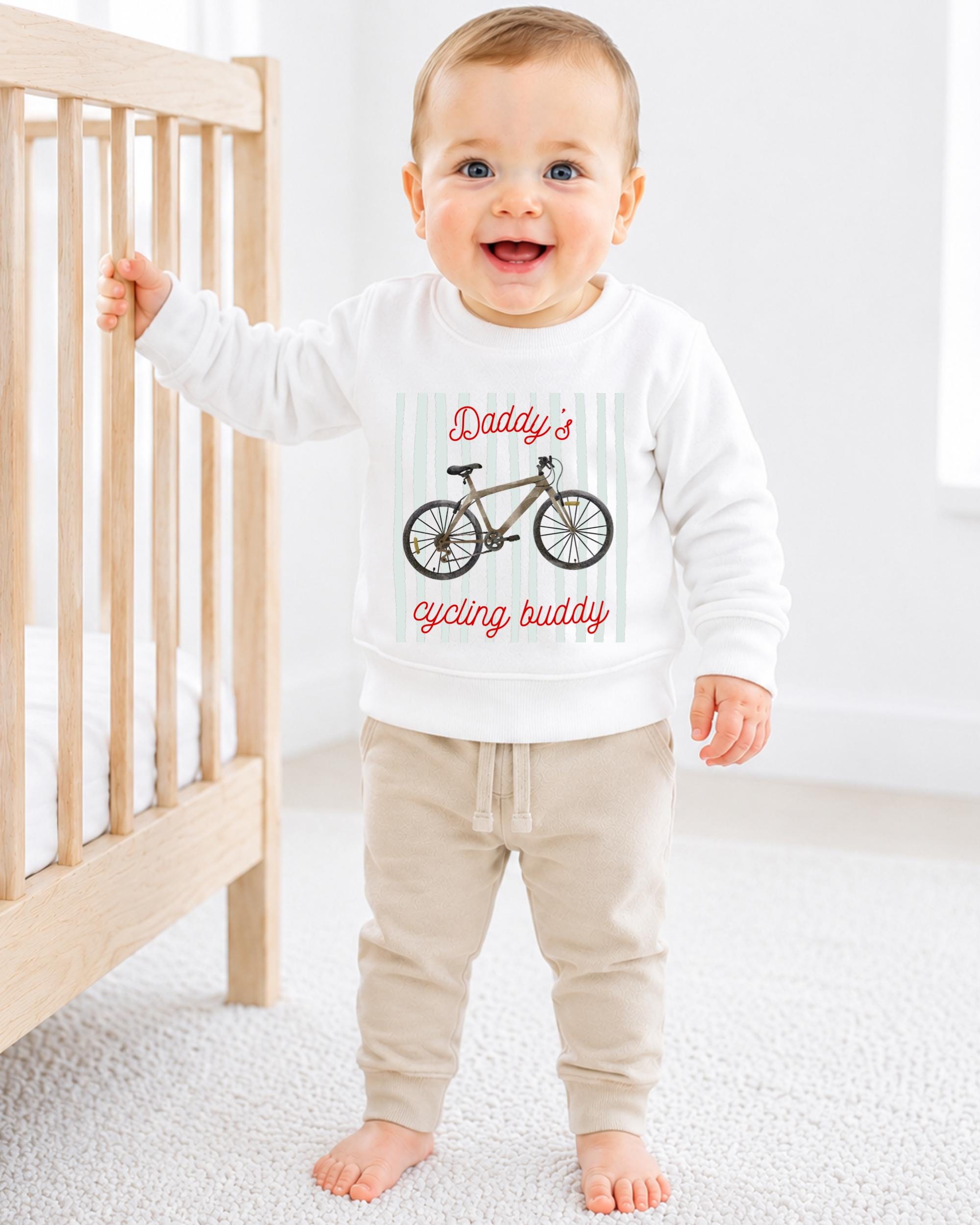 A smiling baby boy wearing a white sweatshirt with a bicycle graphic and the text "Daddy's cycling buddy" is standing next to a wooden crib.