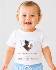A smiling baby wearing a white t-shirt with a heart-shaped photo of a man holding a baby, along with text that reads "Happy 1st Birthday as my Daddy Love you lots, Olivia X".