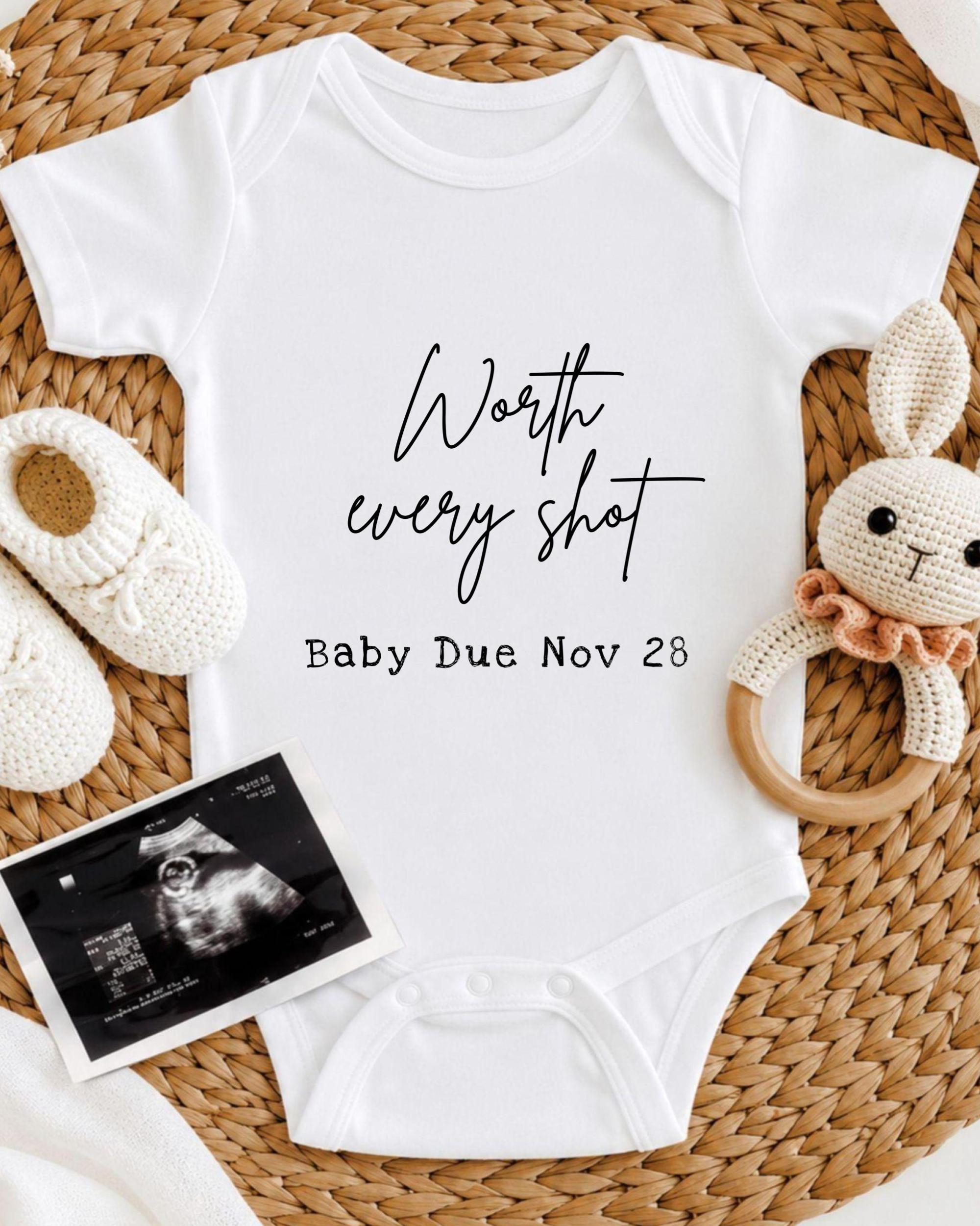 A white baby onesie with the text " Worth every shot" and "Baby Due Nov 28" printed on it, along with a crocheted bunny toy and a baby photograph.