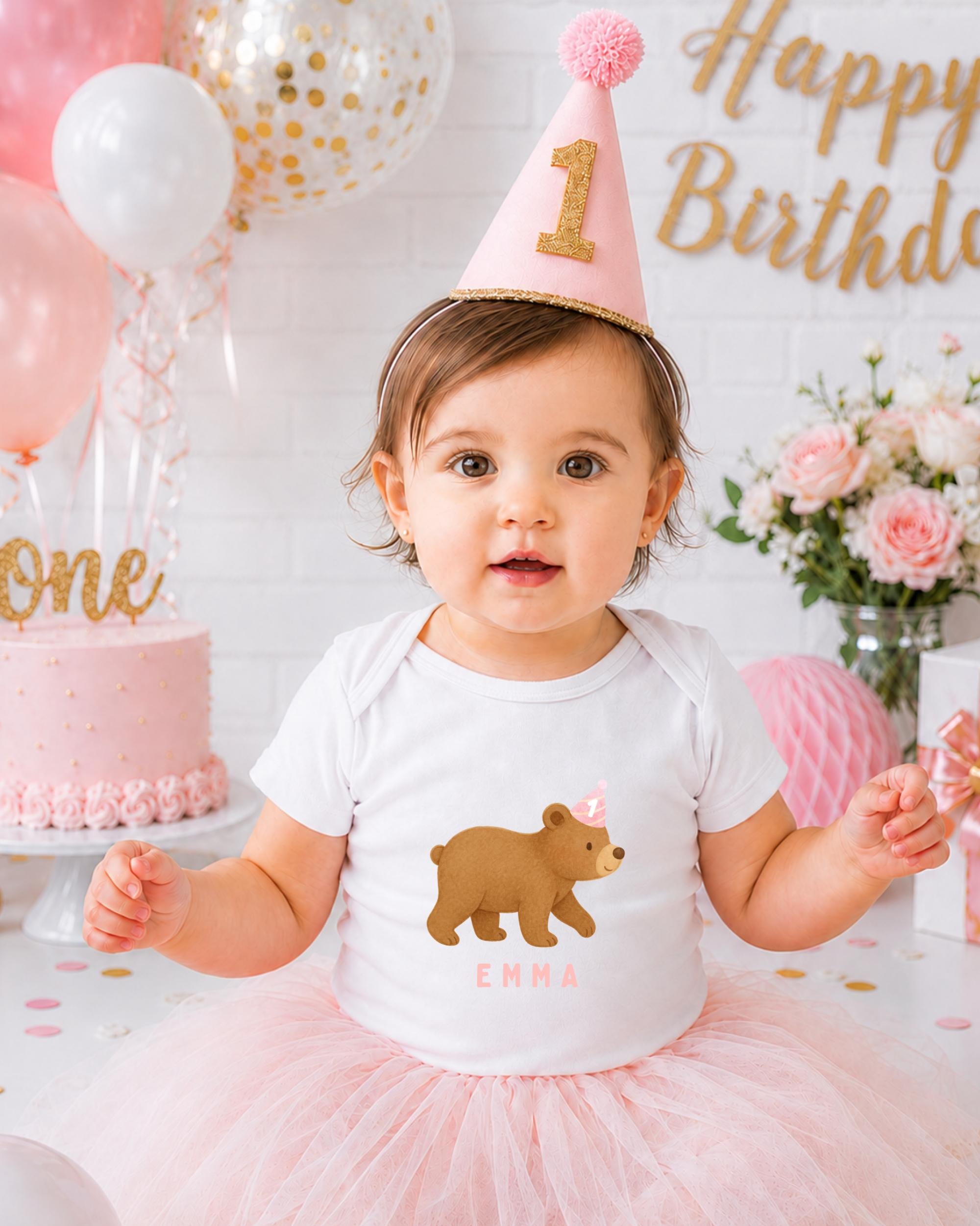 A young girl wearing a birthday hat and a white t-shirt with a bear on it, surrounded by pink and white decorations, including balloons and a cake.