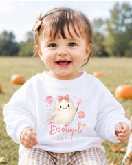 A smiling baby girl wearing a white sweater with a cartoon ghost and the text "Little Bootful Annie" is sitting in a field with pumpkins in the background.