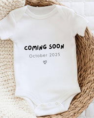 A white onesie with the text "COMING SOON October 2025" printed on it, placed on a woven basket.