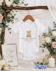 A white baby onesie with a teddy bear design hangs on a wooden hanger, surrounded by floral decorations and a sign that reads "Page Boy".
