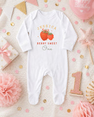 A white onesie with a strawberry design and the text "JESSICA BERRY SWEET One" printed on it, along with a pink star and a pink and gold confetti-covered background.