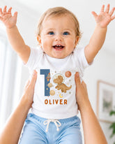 A smiling baby wearing a white bodysuit with a dinosaur and space-themed design, with the name "Olivier" printed on it.