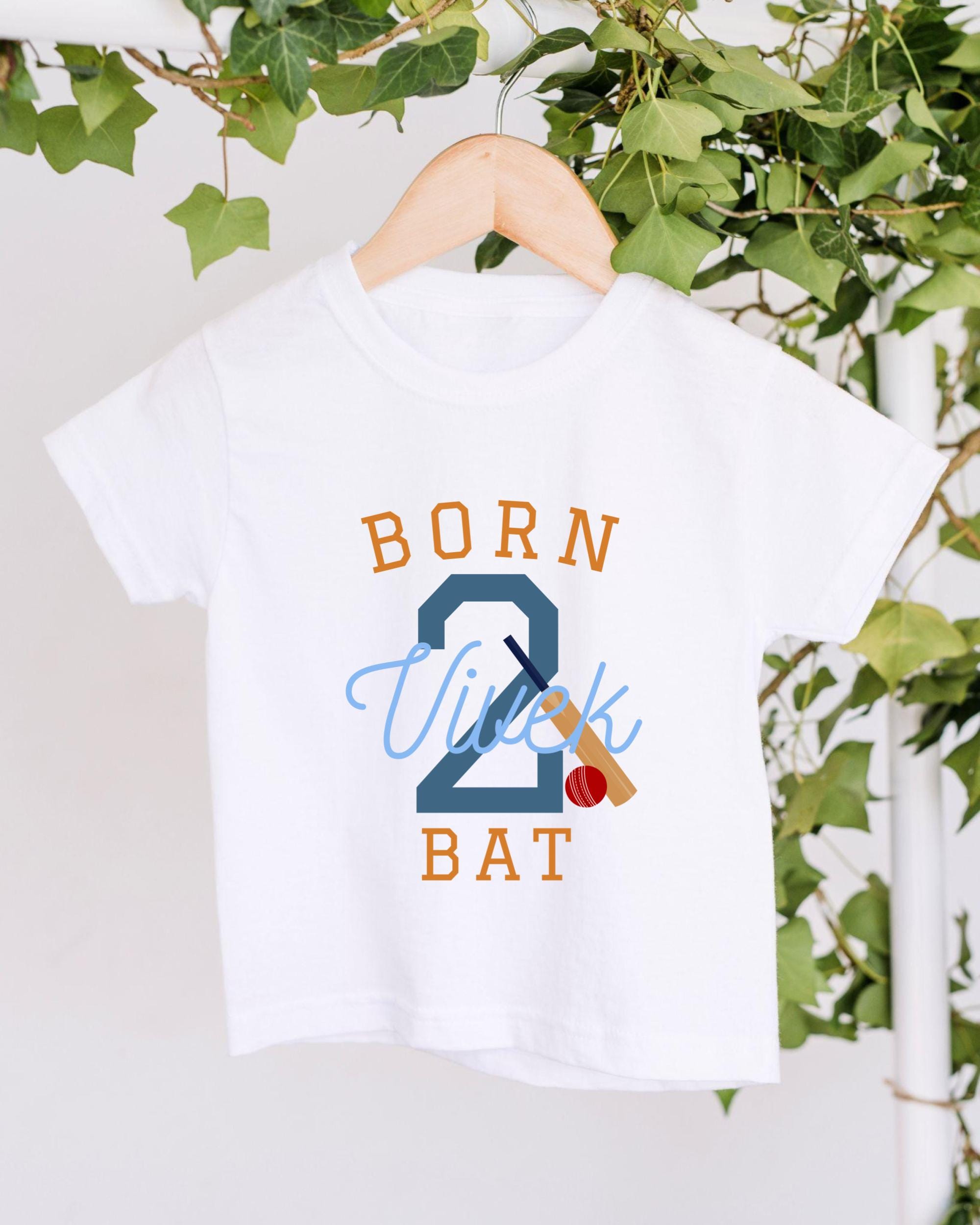 A white t-shirt with the text "BORN 2 VUITK BAT" printed on it, along with a cricket bat and ball graphic.