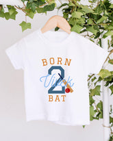 A white t-shirt with the text "BORN 2 VUITK BAT" printed on it, along with a cricket bat and ball graphic.