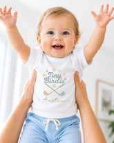 A smiling baby wearing a white t-shirt with a graphic design and the name "Tiny Birdie" printed on it, being held up by someone's arms.