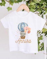 A white t-shirt with a printed design of a gray elephant in a basket, surrounded by colorful balloons, and the name "Annisa" written below.