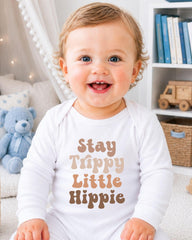 A smiling baby wearing a white onesie with the text "Stay Trippy Little Hippie" printed on it.