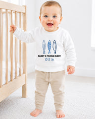 A smiling baby boy wearing a white sweatshirt with a design of fish and the text "DADDY'S FISHING BUDDY OLLIE" is standing next to a wooden crib.
