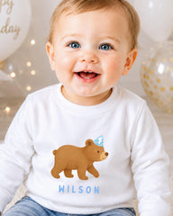 A smiling baby boy wearing a white shirt with a cartoon bear and the name "Wilson" printed on it.