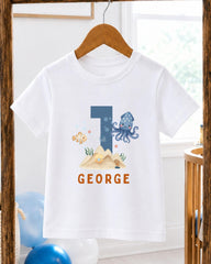 A white t-shirt with a graphic design featuring a blue octopus, a goldfish, and a sand castle, along with the text "GEORGE" printed on it.