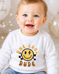 A smiling baby boy wearing a white shirt with a smiley face and the text "One cool dude" printed on it.