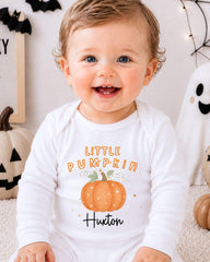 A smiling baby boy wearing a white onesie with a pumpkin design and the text "Little Pumpkin" is sitting in front of Halloween decorations.