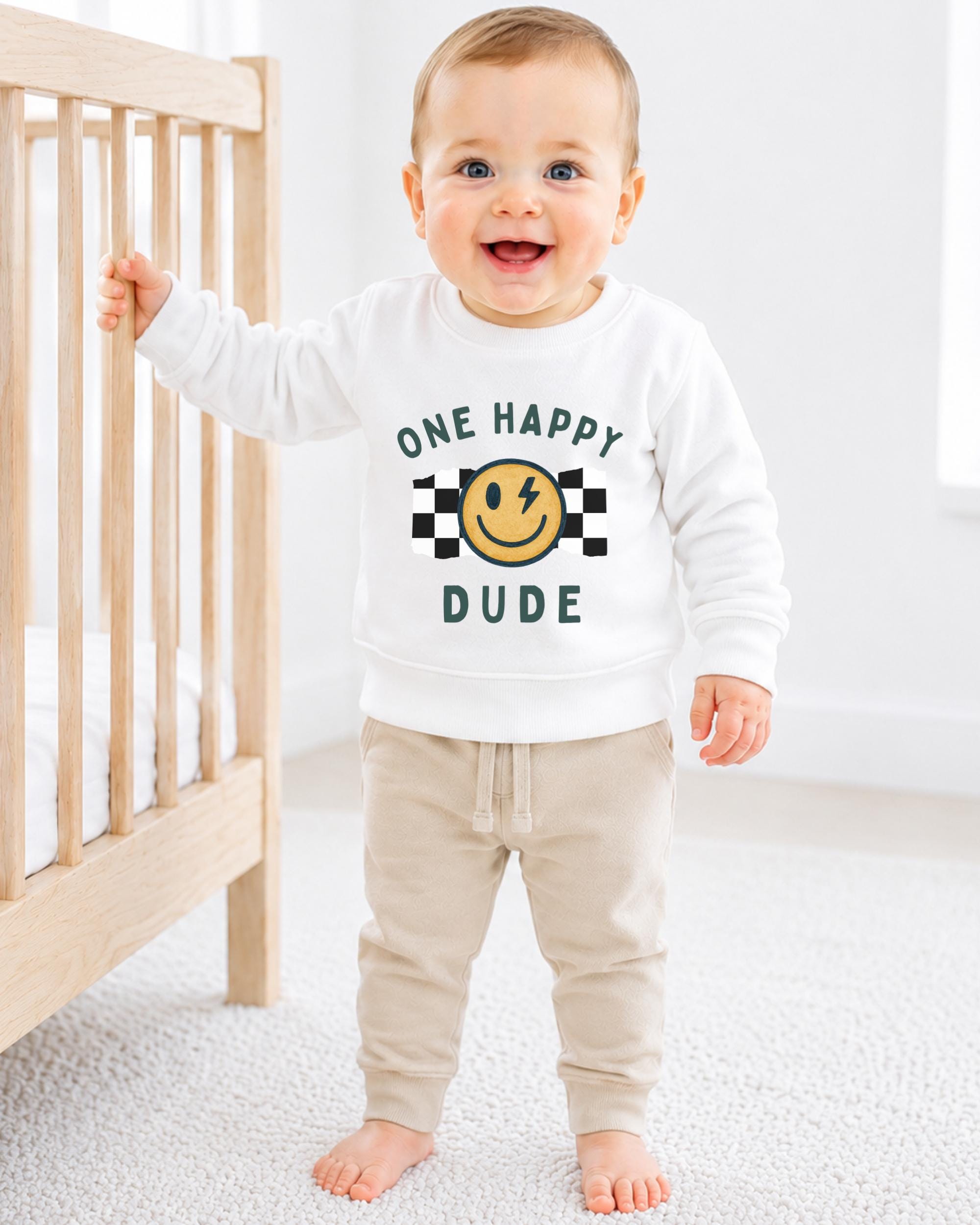 A smiling baby boy wearing a white sweatshirt with a smiley face and the text "One happy dude" is standing next to a wooden crib.