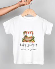 A white t-shirt with a graphic design of a fruit and vegetable stand, along with the text "Baby Jackson Locally grown".