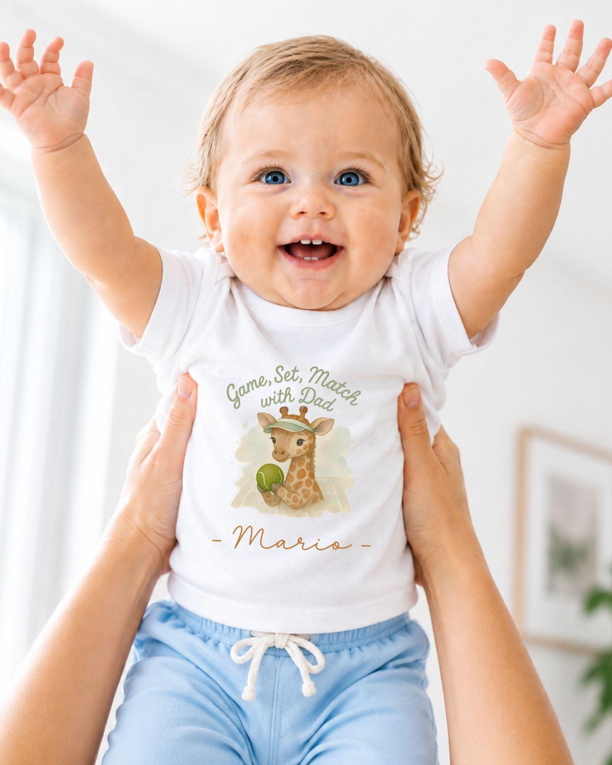 A smiling baby wearing a white t-shirt with a giraffe graphic and the text "Game, Set, Match with Dad - Mario" is being held up by someone's arms.