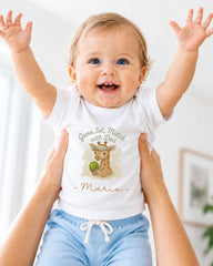 A smiling baby wearing a white t-shirt with a giraffe graphic and the text "Game, Set, Match with Dad - Mario" is being held up by someone's arms.