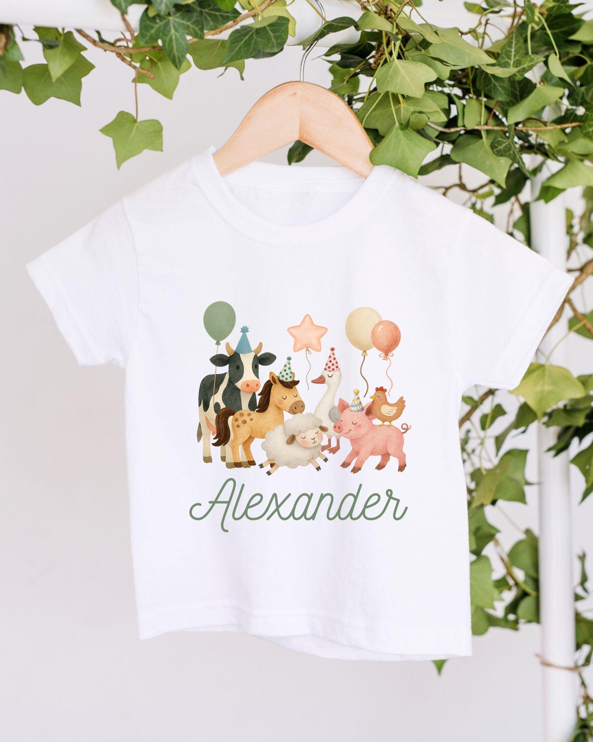A white t-shirt with a colorful illustration of various animals, including cows, horses, and a pig, along with balloons and a star. The name "Alexander" is written below the illustration.