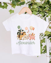 A white t-shirt with a colorful illustration of various animals, including cows, horses, and a pig, along with balloons and a star. The name "Alexander" is written below the illustration.