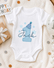 A white onesie with a blue number one and stars, along with a "Happy Birthday Jack" sign, is displayed on a white blanket.