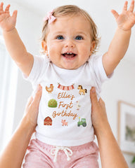 A smiling baby girl wearing a white t-shirt with farm animal illustrations and the text "Ellie's First Birthday" is being held up by someone's arms.