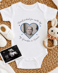 A white baby onesie with a photo of an older man inside a heart shape, surrounded by a crocheted bunny toy and a baby bootie.