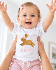 A smiling baby girl wearing a white t-shirt with a bunny graphic and the name "EMILY" printed on it, being held up by someone's arms.