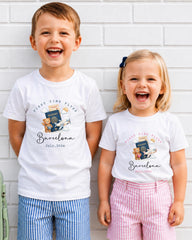 Two children, a boy and a girl, are standing next to each other and smiling, wearing matching white t-shirts with a graphic design and text.