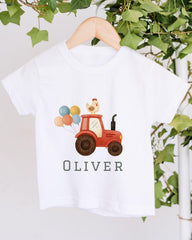 A white t-shirt with a graphic design of a red tractor carrying colorful balloons and a chicken on top, along with the name "Olivier" printed below.