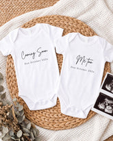 Two white baby onesies with text that announces the upcoming arrival of a baby, placed on a woven placemat.