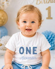 A smiling baby boy wearing a white t-shirt with the text "ONE Alexander" and blue shorts, sitting in front of a birthday party backdrop with balloons and confetti.