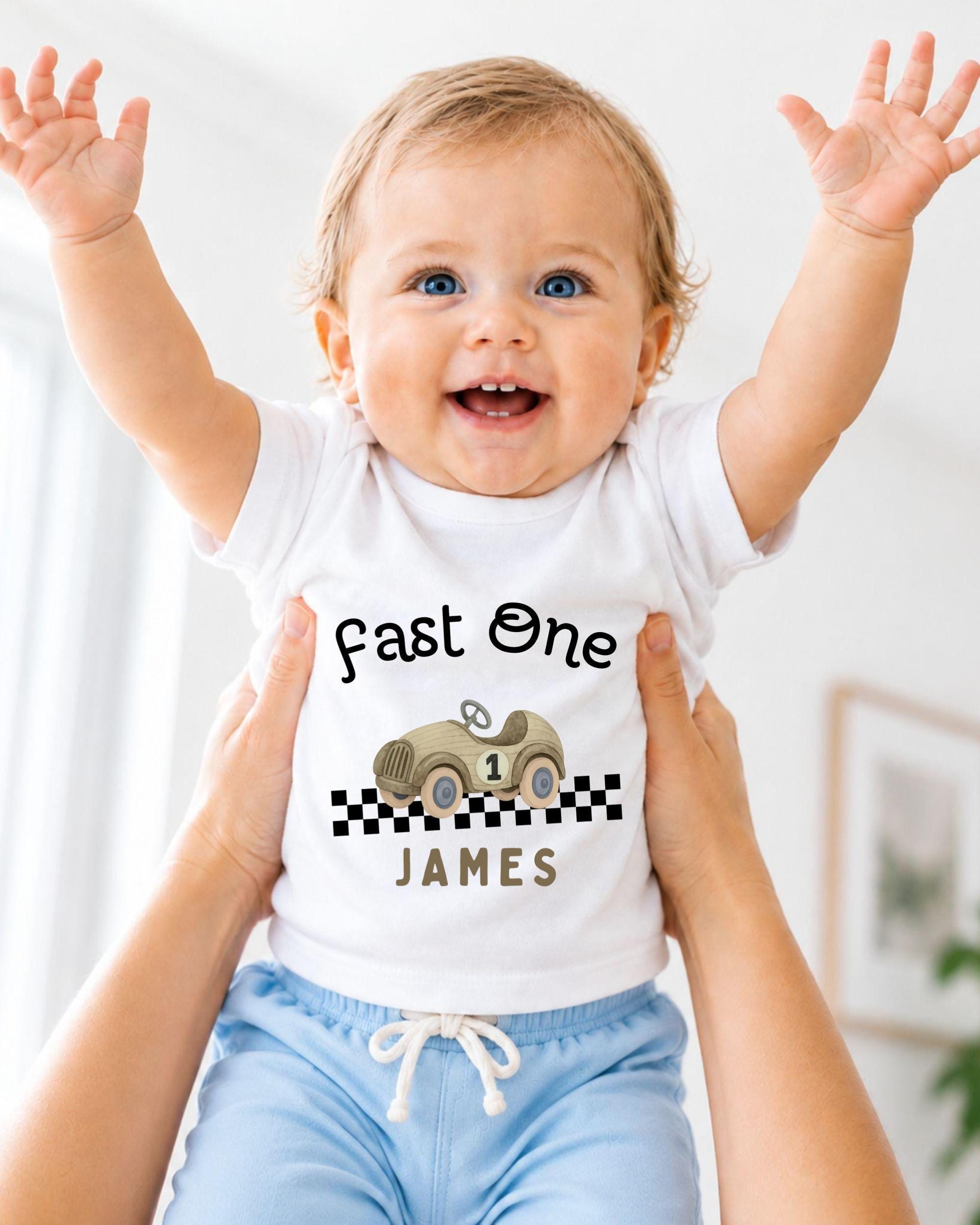A smiling baby wearing a white t-shirt with a race car graphic and the text "Fast One James" is being held up by someone's arms.