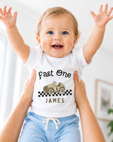 A smiling baby wearing a white t-shirt with a race car graphic and the text "Fast One James" is being held up by someone's arms.