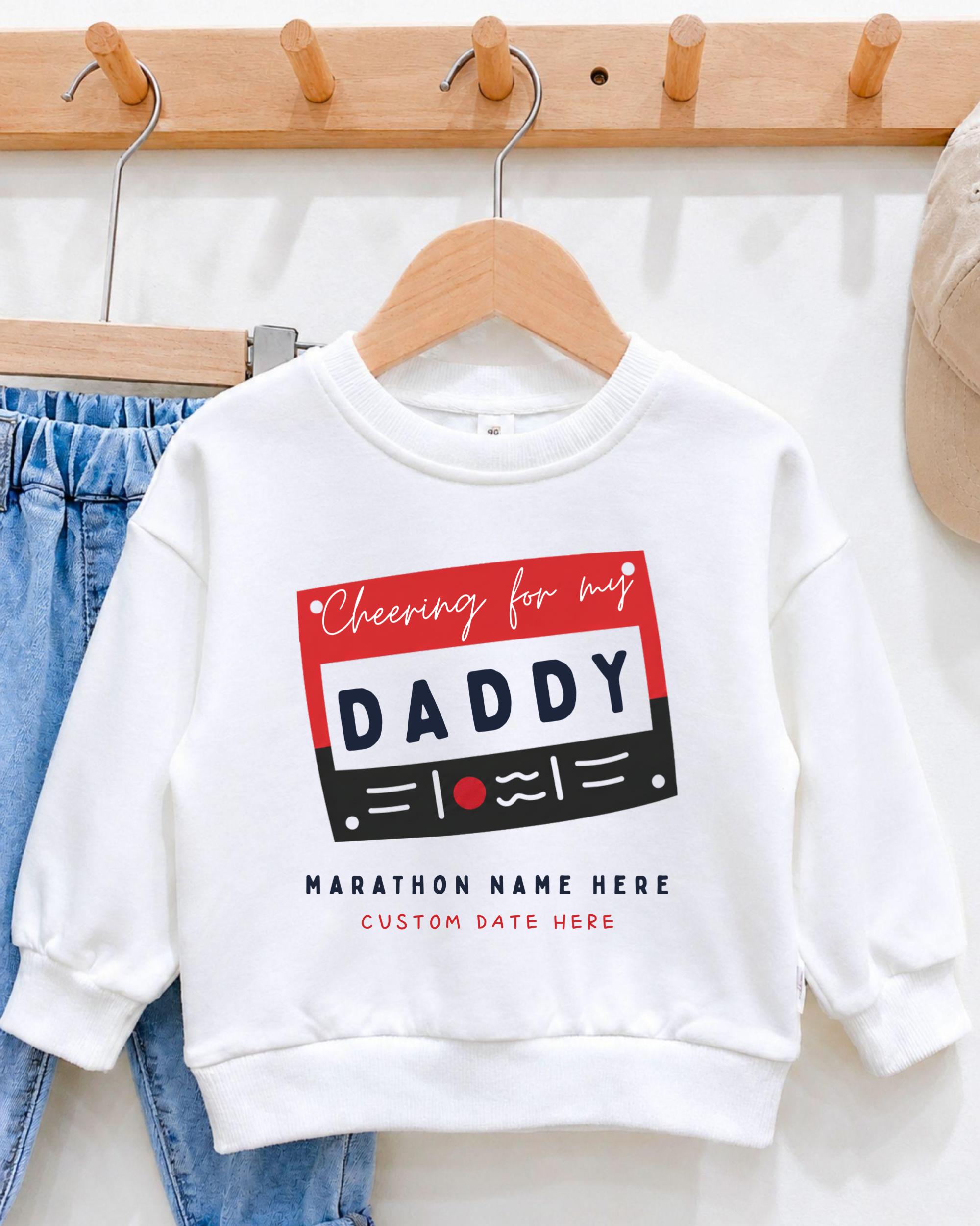 A white sweatshirt with a graphic design that says "Cheering for my DADDY" printed on it, hanging on a wooden hanger.