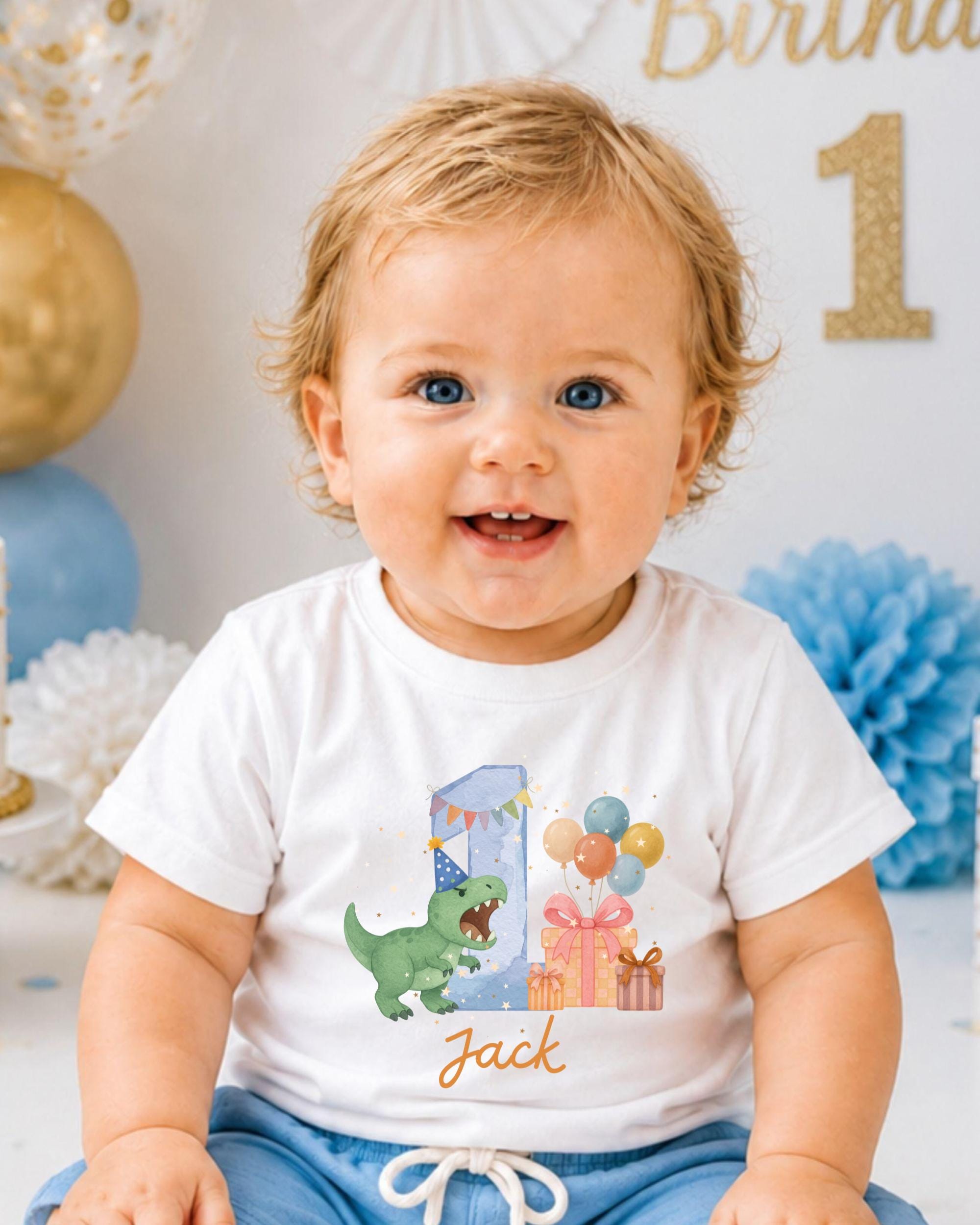 A smiling baby boy wearing a white t-shirt with a dinosaur graphic and birthday decorations.