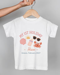 A white t-shirt with a graphic design featuring a baby bottle, sunglasses, a crab, and a sun, along with the text "MY 1ST HOLIDAY Alice - Greece, February 2027".