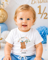 A smiling baby boy wearing a white t-shirt with a cartoon bear and text, sitting in front of a birthday party decoration with gold and blue balloons.