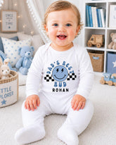 A smiling baby boy wearing a white onesie with a graphic and text, sitting on a carpeted floor in a room with a bookshelf and stuffed animals in the background.