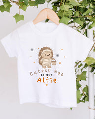 A white t-shirt with a cartoon hedgehog wearing a mummy costume and the text "Cute in town" printed on it, hanging on a wooden hanger.