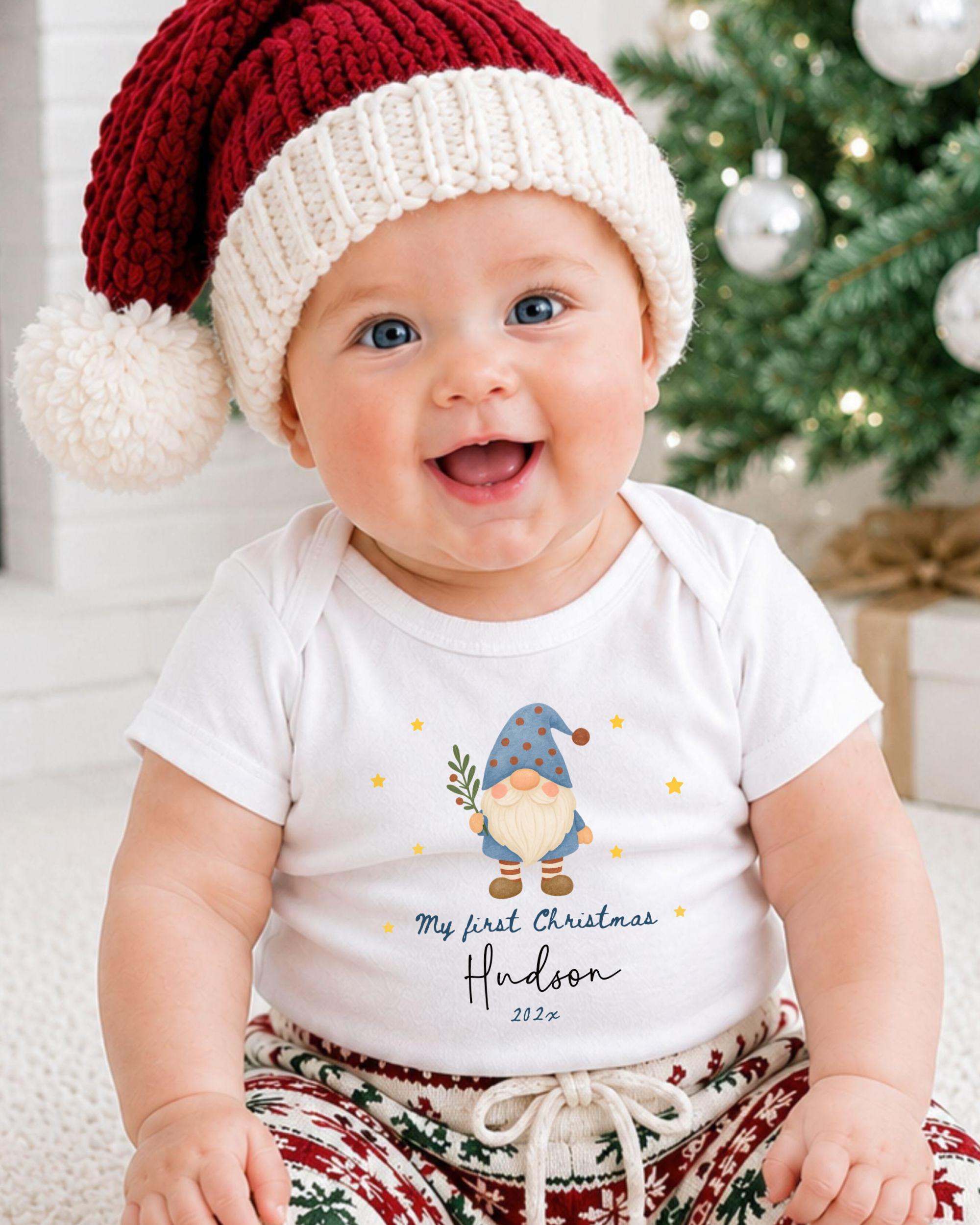 A smiling baby wearing a Santa hat and a white t-shirt with a gnome graphic, sitting in front of a Christmas tree.