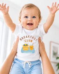 A smiling baby wearing a white shirt with a breakfast club graphic and blue shorts, being held up by someone's arms.