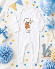 A white onesie with a cartoon bear and the text "Half way to one" is displayed among various baby-related items, including a blue balloon, confetti, and a star decoration.