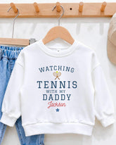 A white sweatshirt with the text "WATCHING TENNIS WITH MY DADDY JACKSON" printed on it, along with a tennis racket graphic.
