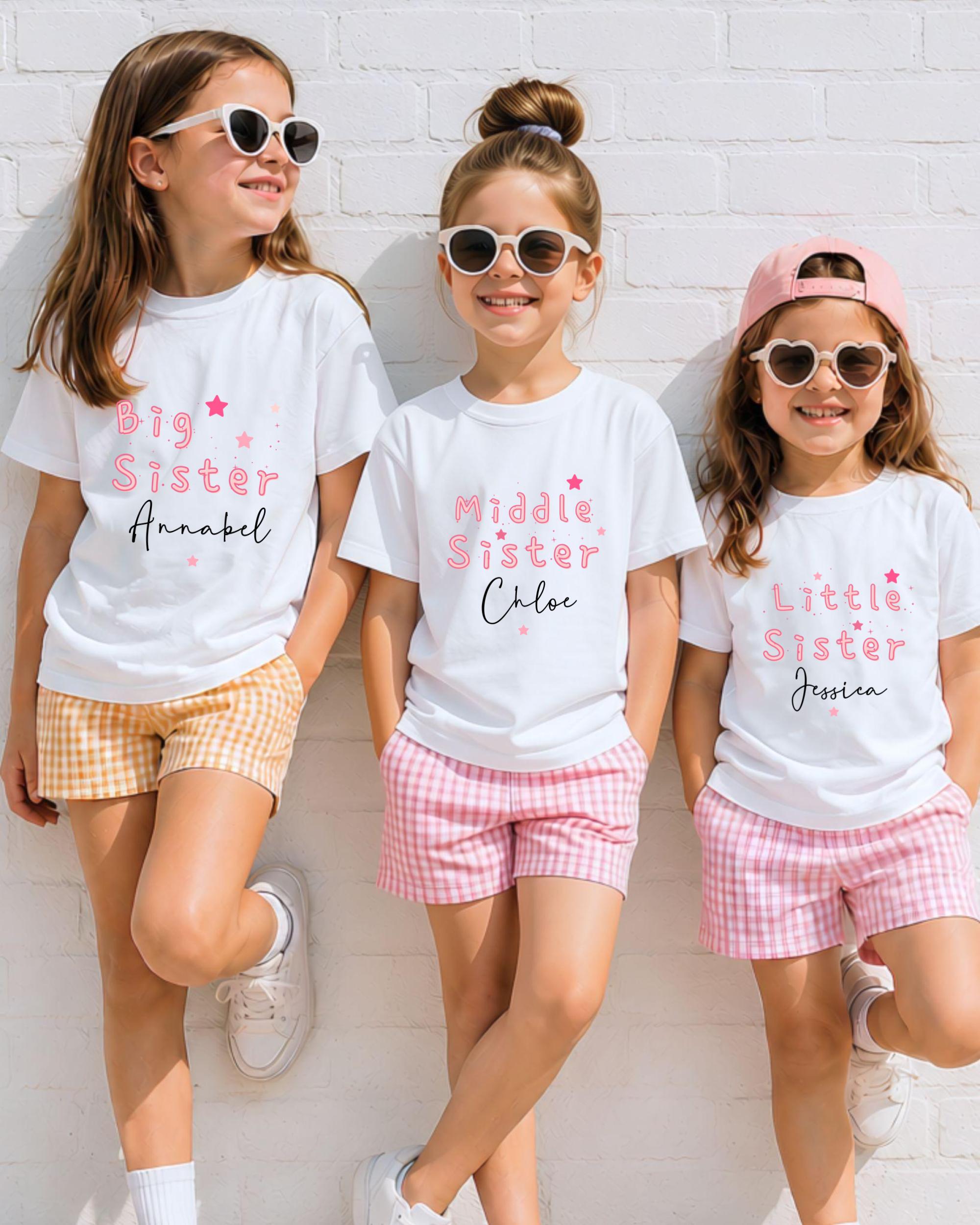 Three young girls wearing white t-shirts with the text "Big Sister" printed on them, along with sunglasses and plaid shorts, standing against a white brick wall.